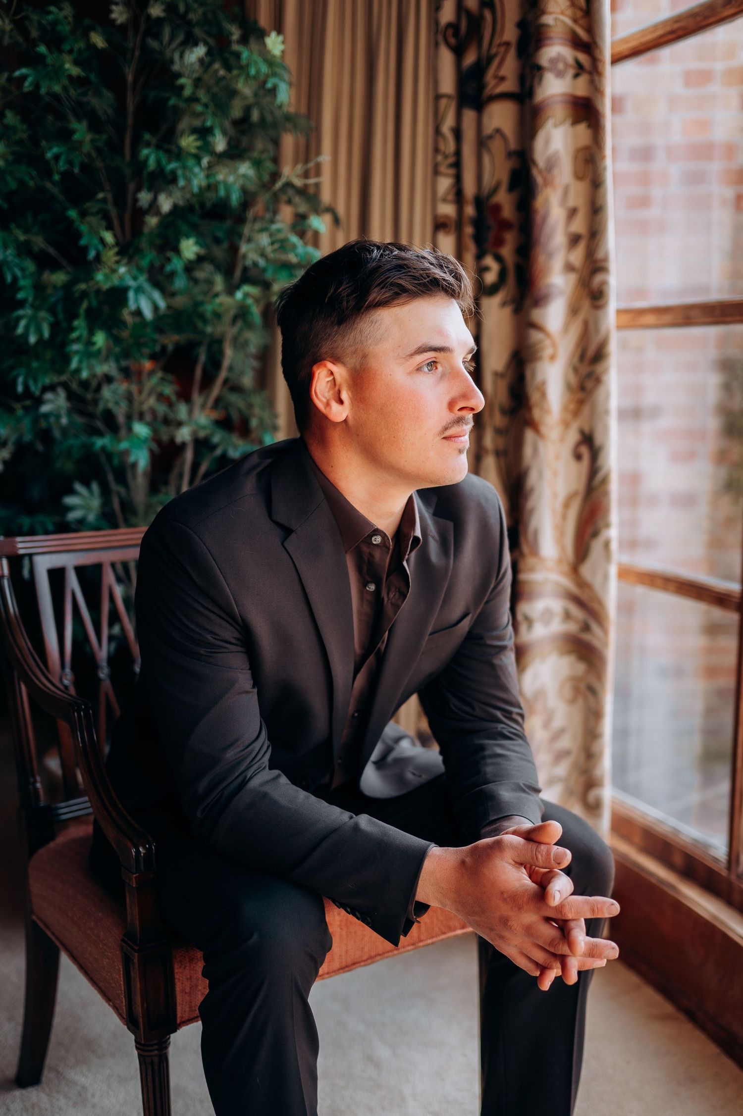 Young man in black suit sitting in wooden chair gazing out window with green plants and curtains in background.