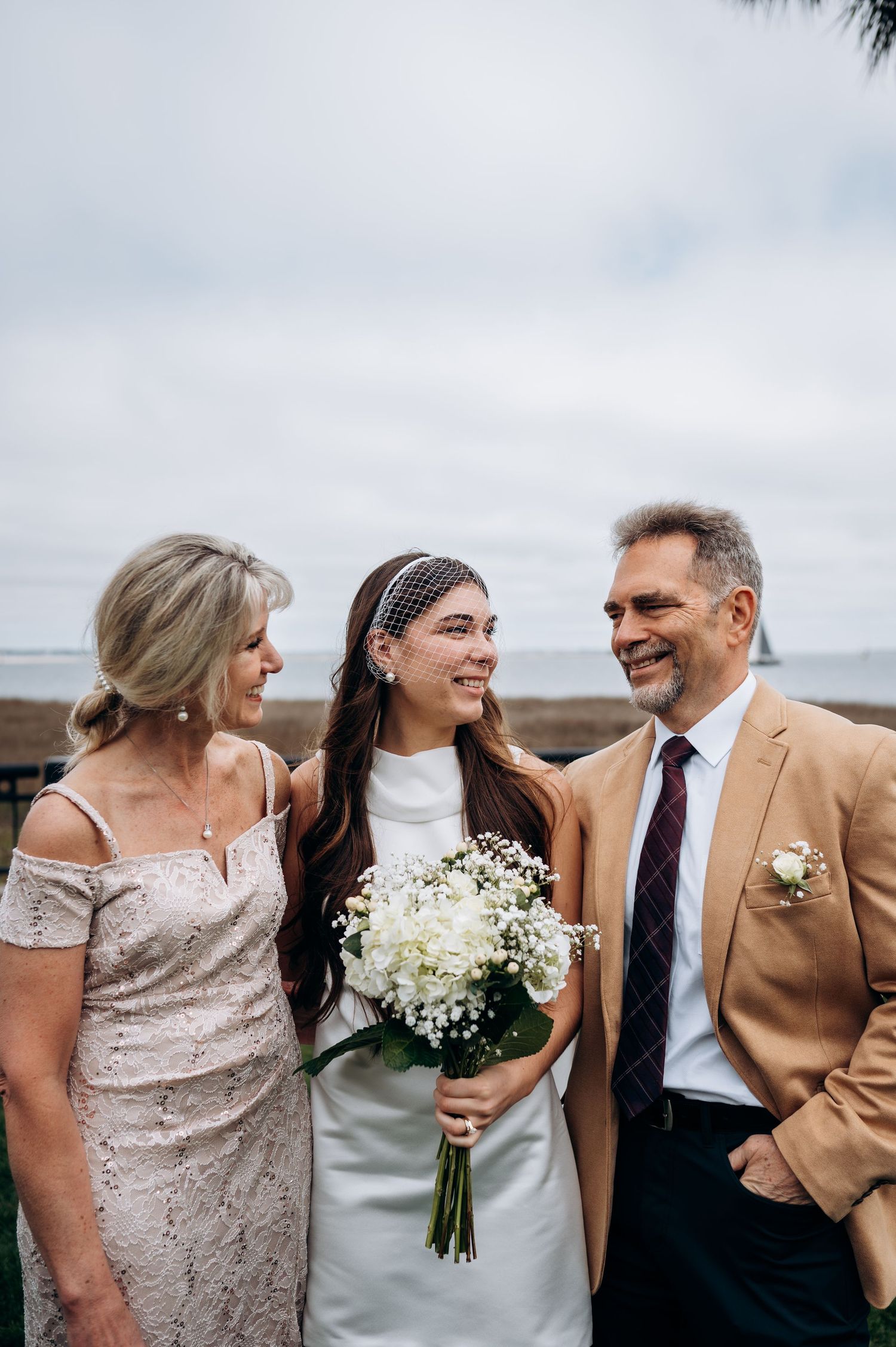 Smiling bride with white bouquet stands between her proud parents on her wedding day by the waterfront.