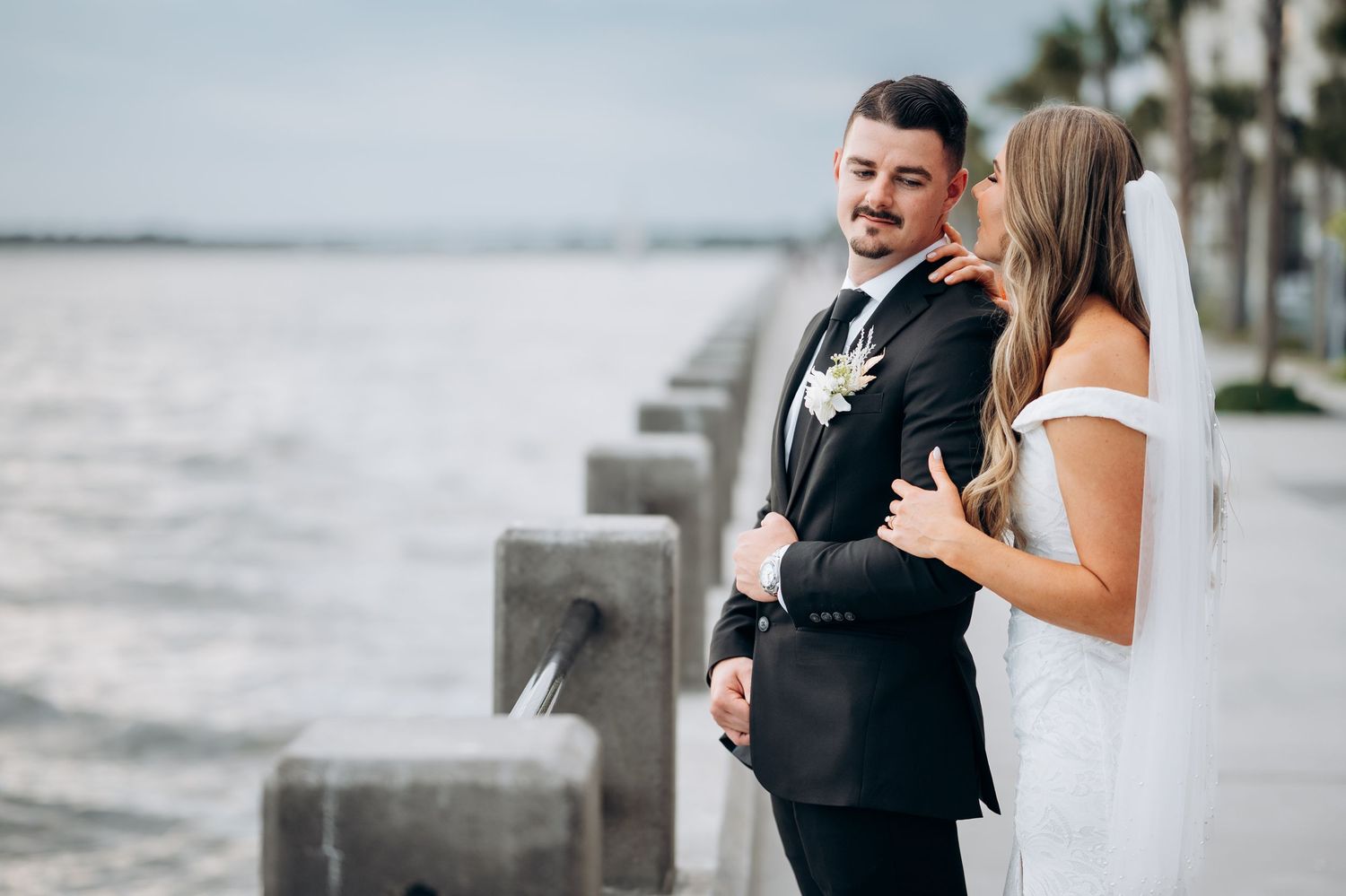 Bride and groom embracing by the waterfront, groom in black suit with white floral boutonniere.