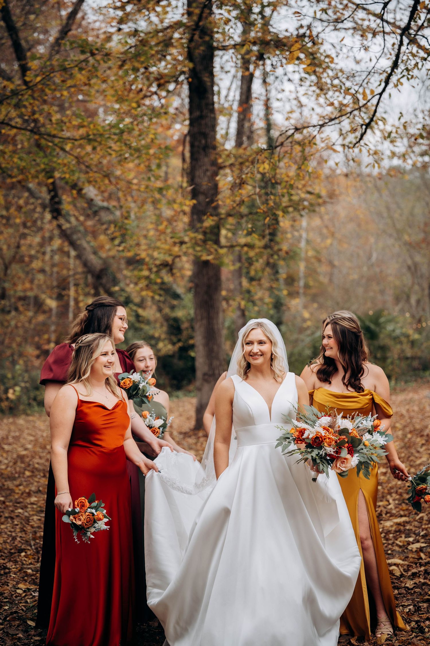 Wedding party laughs together while walking through autumn leaves in burgundy and white attire.