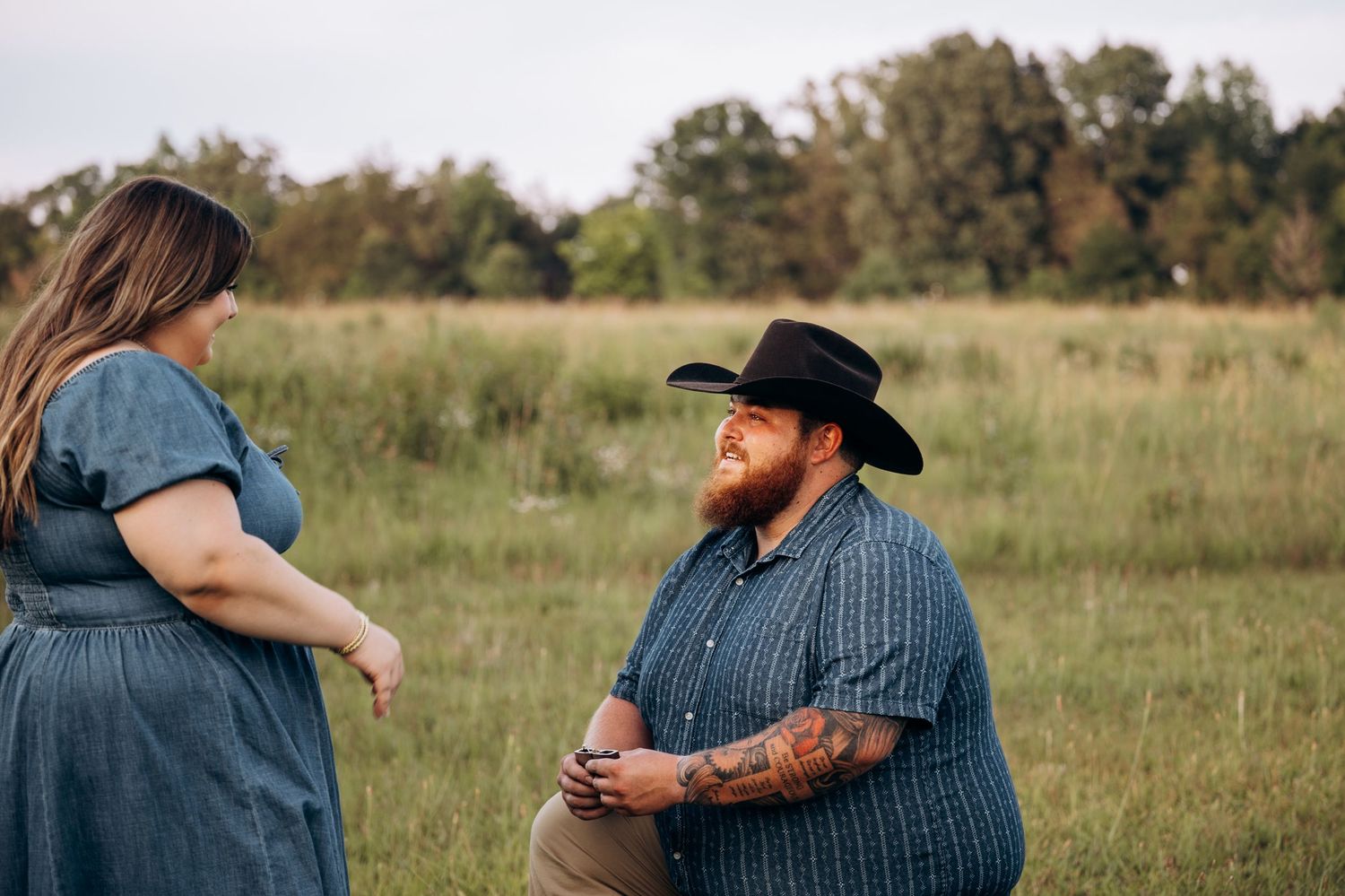 Bearded man in black cowboy hat kneeling in a green field during a marriage proposal.