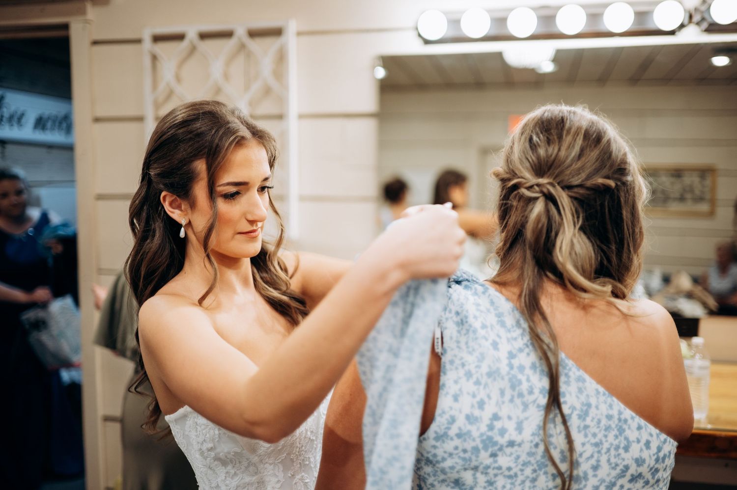 Bride helping bridesmaid zip up blue floral dress in a dressing room before wedding ceremony.