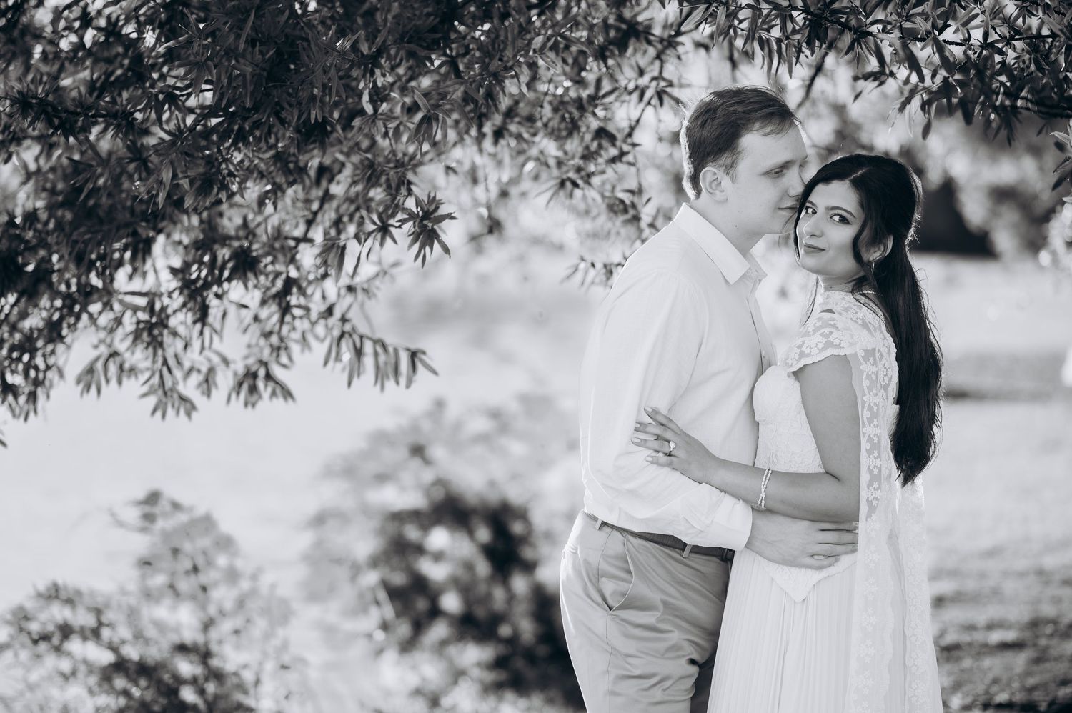 Black and white photo of couple embracing tenderly under willow tree branches in outdoor setting.