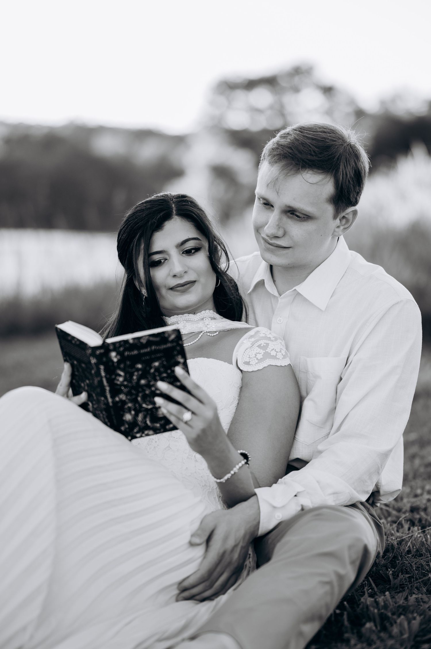 Black and white photo of a couple sitting outdoors, reading a book together in wedding attire.