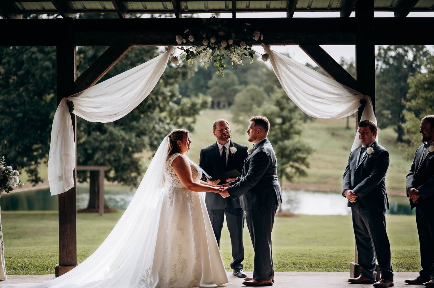 Wedding ceremony setup under a covered outdoor pavilion with white draping and natural scenery.