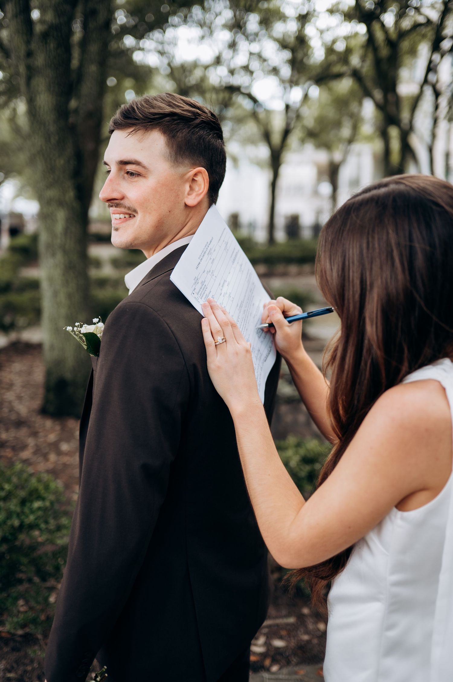 A smiling groom in a dark suit uses his back as a desk while a bride in white signs their marriage certificate outdoors.