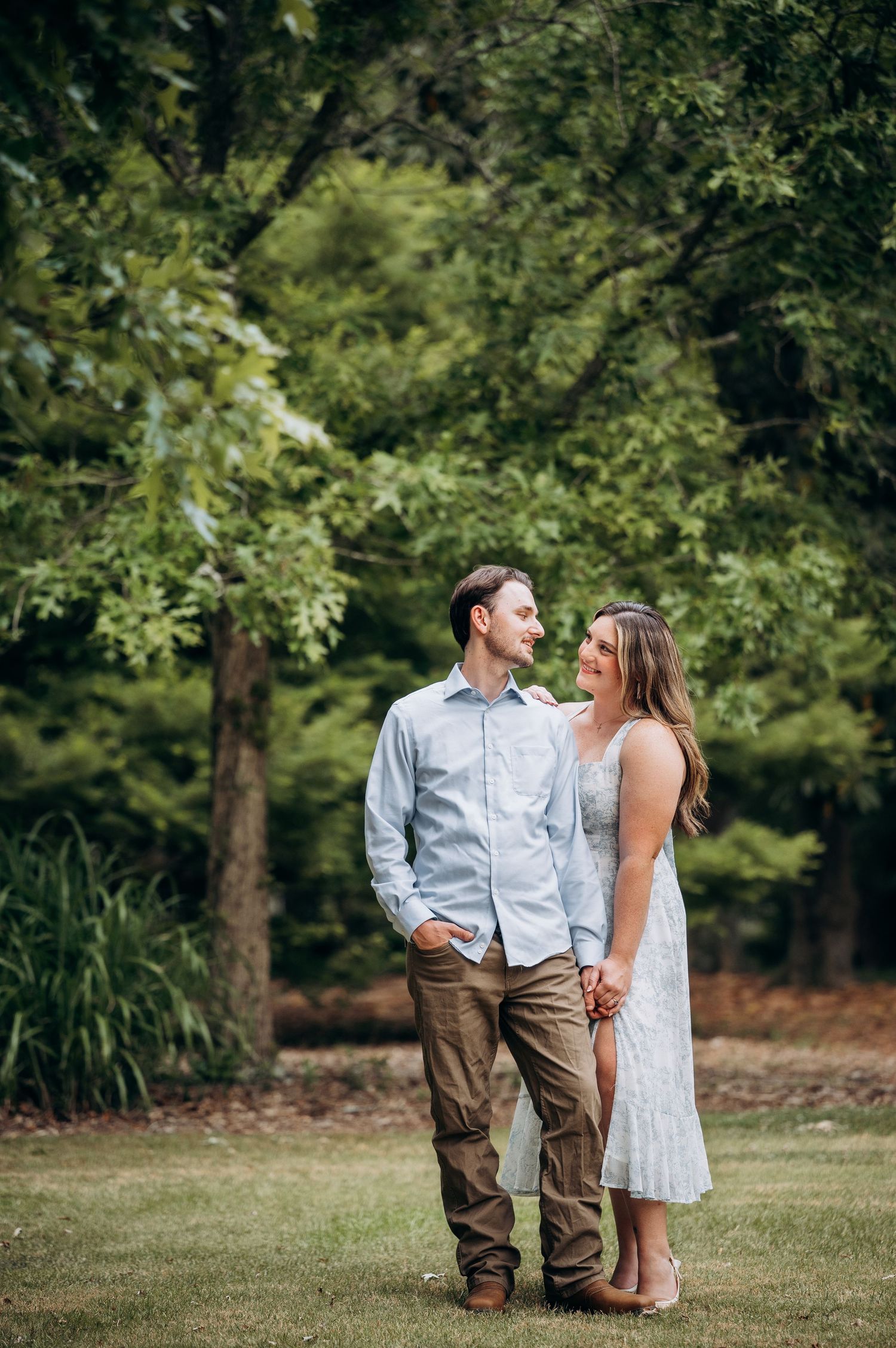 Couple standing together on green lawn surrounded by lush trees, man in blue shirt and woman in white dress smiling at each other.