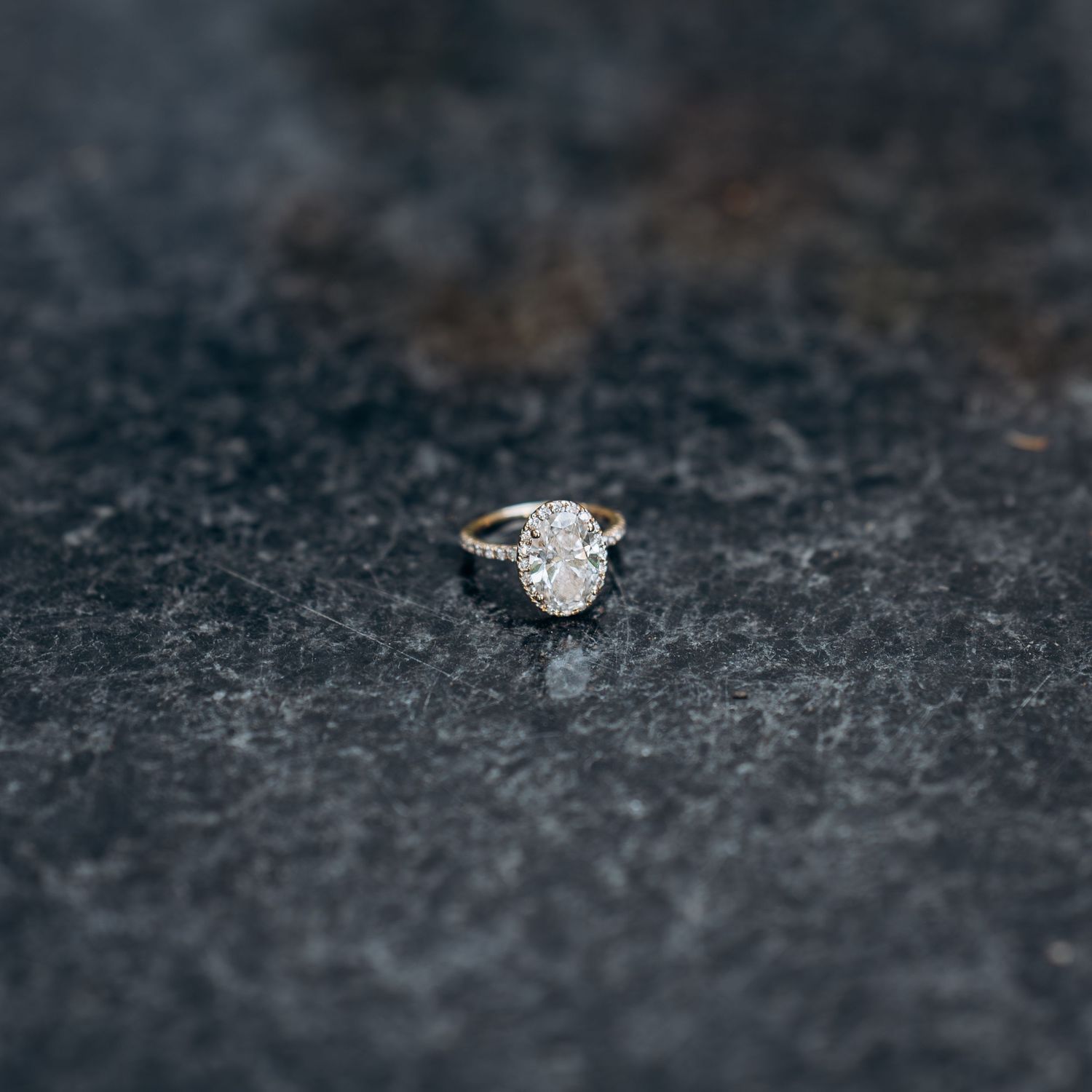 Elegant oval diamond engagement ring with halo setting displayed on dark textured stone surface.