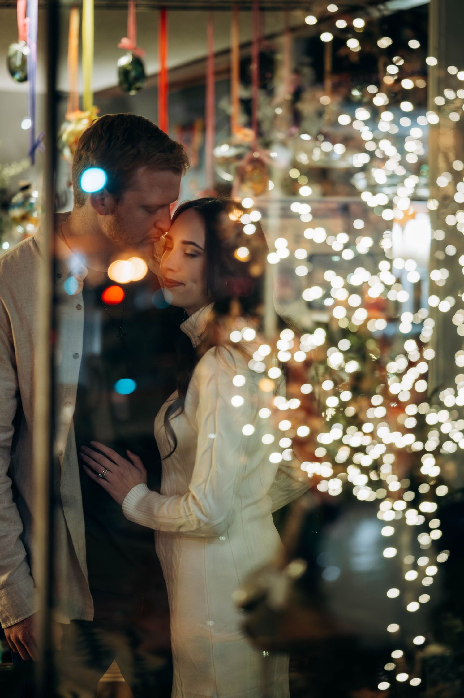 Couple sharing a tender kiss surrounded by sparkling Christmas tree lights during a festive pregnancy announcement shoot.