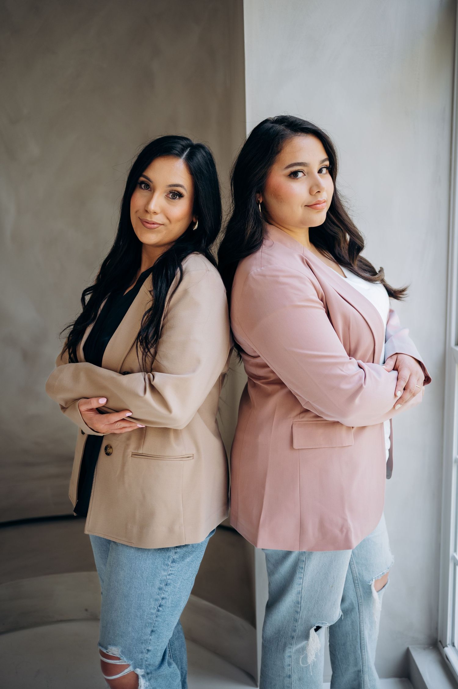 Two women in casual attire pose back to back in a modern interior setting.