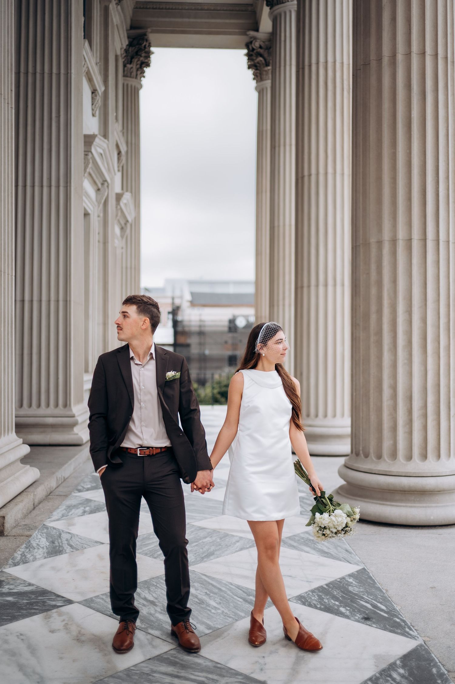 Romantic couple poses between classic stone columns in formal attire with white flowers.