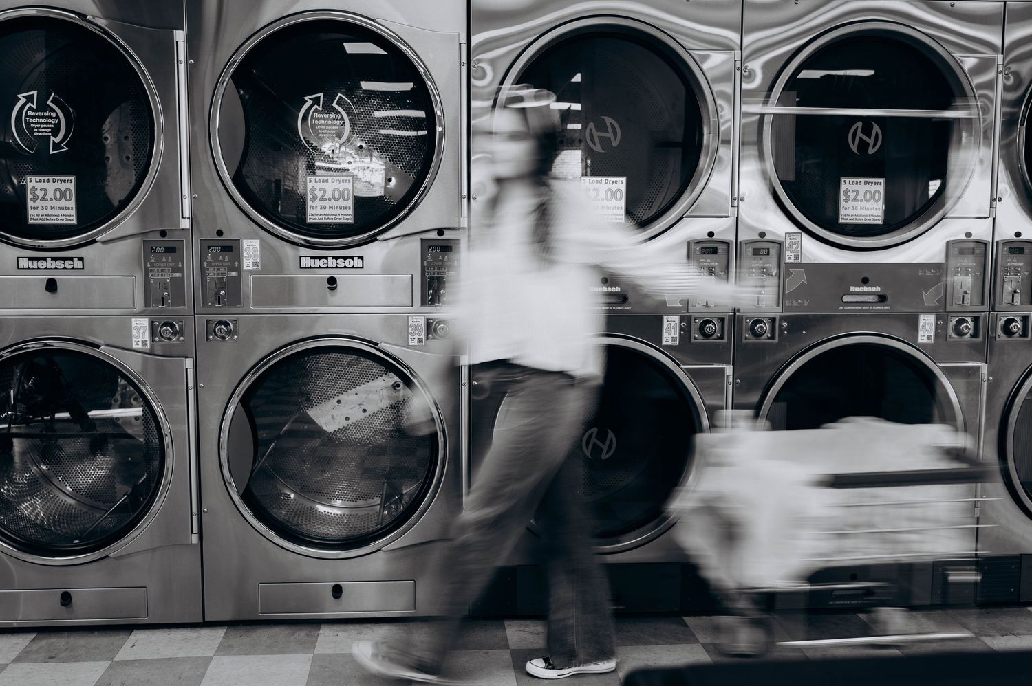 Row of industrial washing machines in a laundromat with chrome doors reflecting light and creating a repeating pattern.