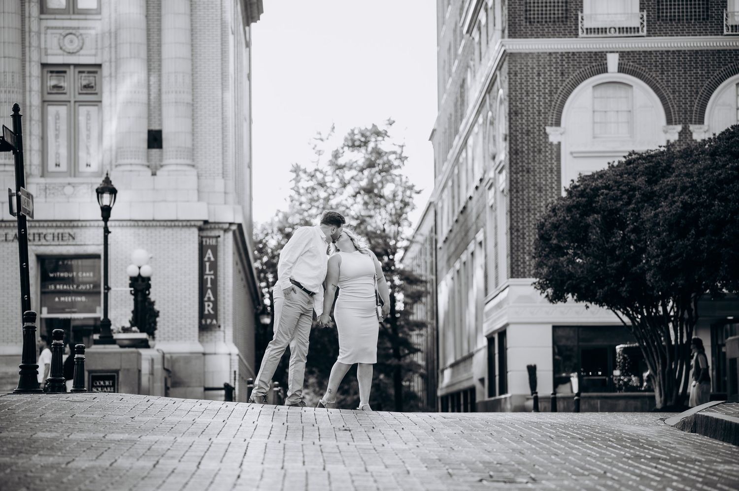 Black and white photo of a couple walking together on a brick plaza surrounded by classic architecture.