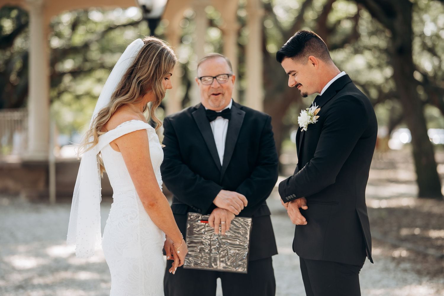 Bride and groom exchanging vows with officiant outdoors under oak trees during elegant wedding ceremony.