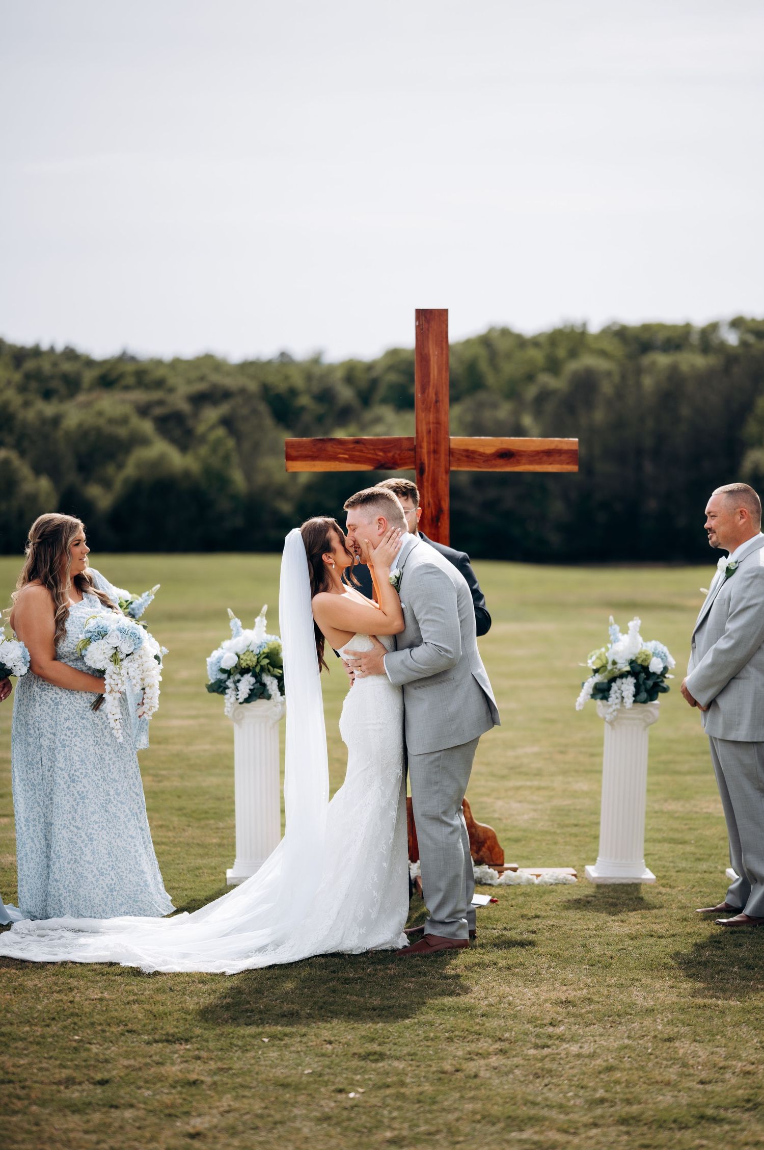 Bride and groom share first kiss at outdoor altar with wooden cross and white floral arrangements.