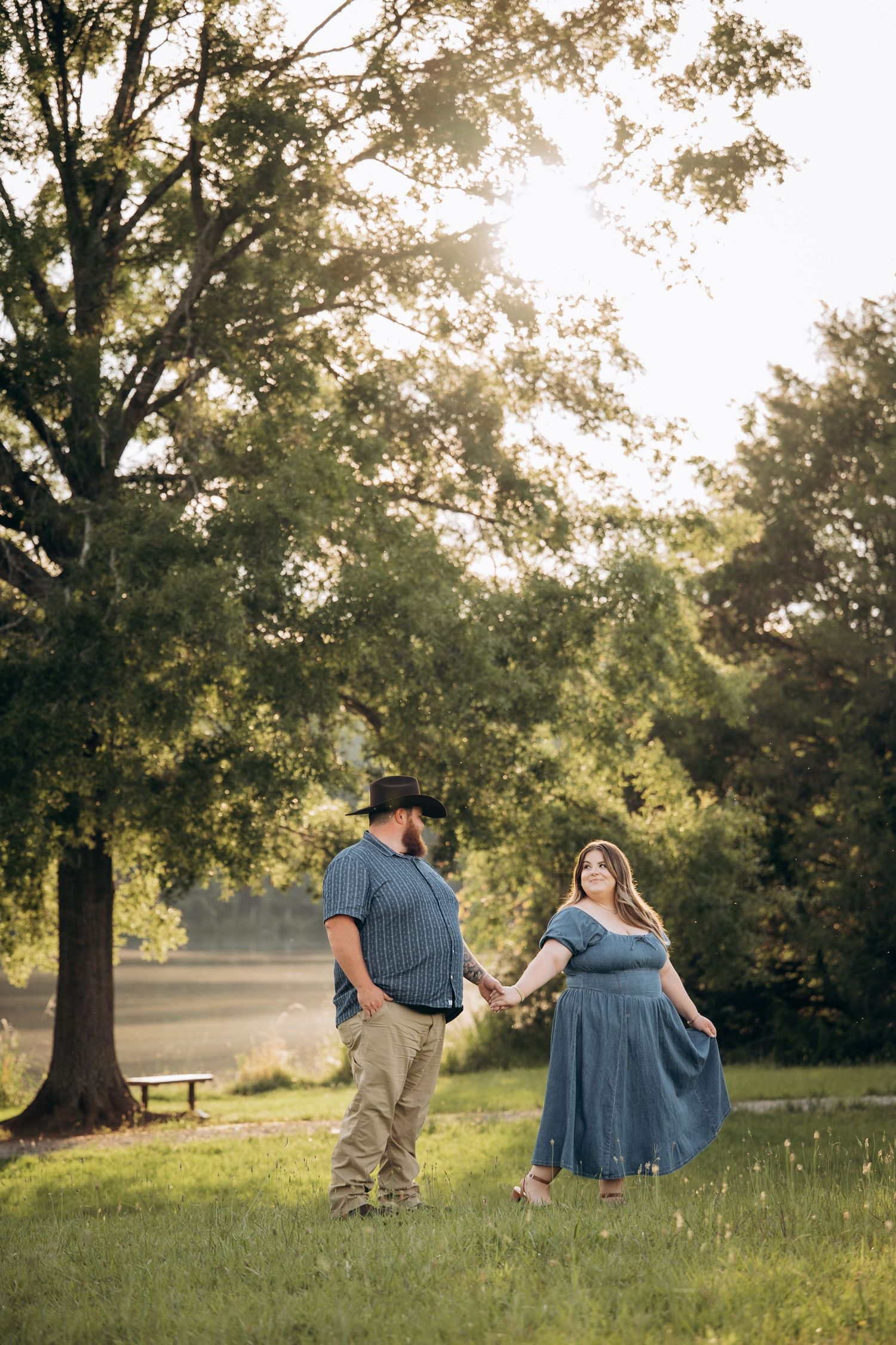 Couple holding hands in a sunlit park, woman in blue dress and man in cowboy hat, golden hour photography.