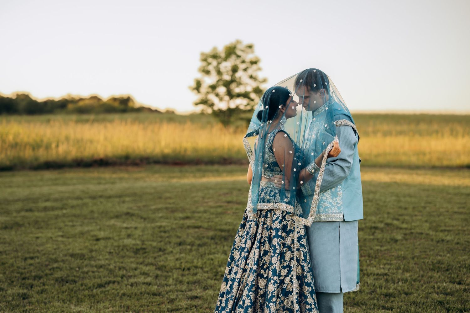 Couple in blue wedding attire sharing romantic moment under veil in golden hour field.