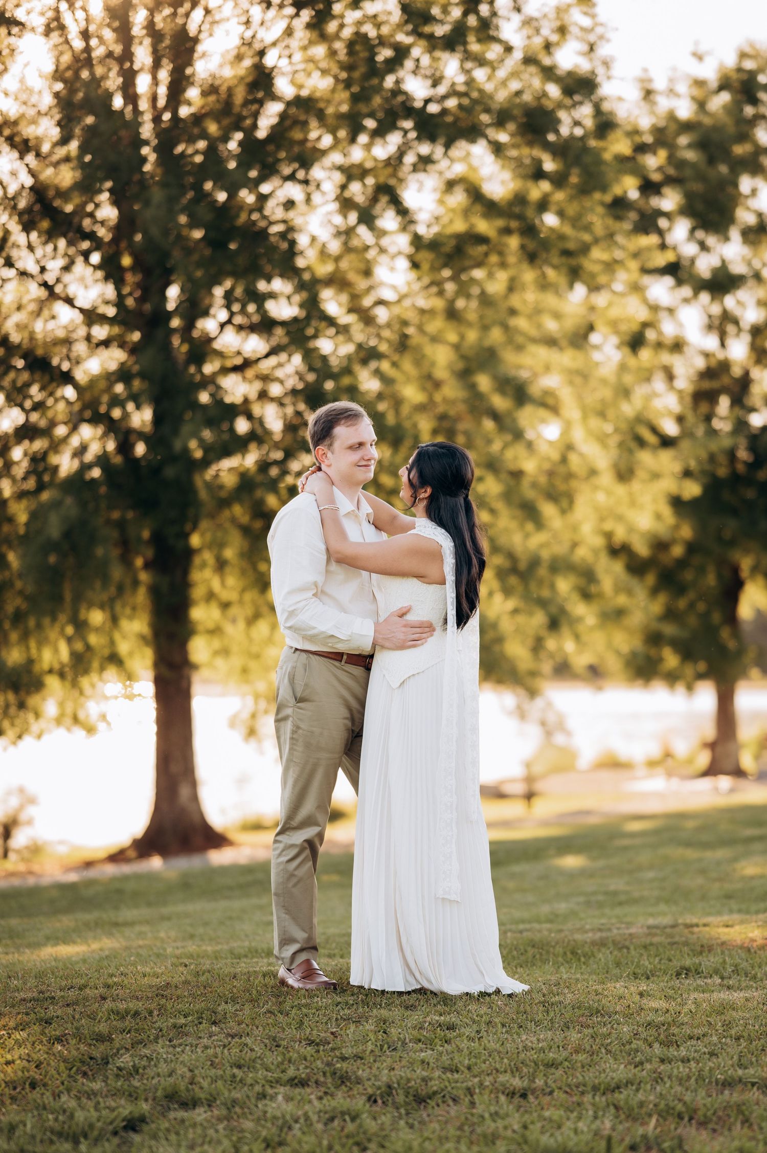 A couple embraces in a sunlit park, the bride in a white gown and groom in khaki, surrounded by lush green trees.