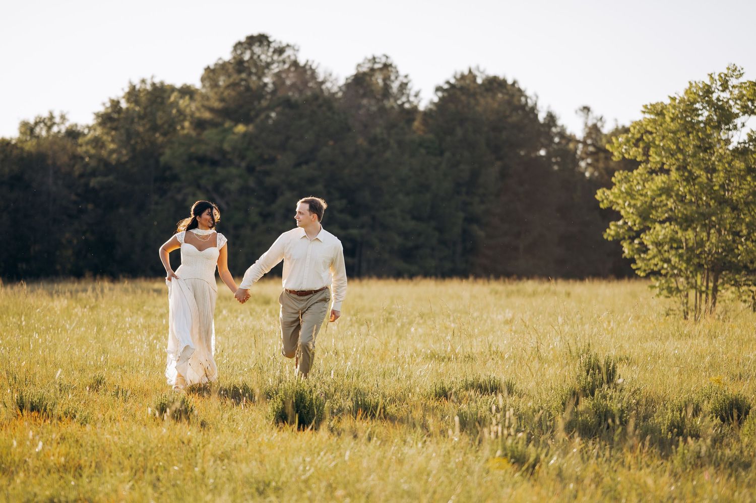 Couple holding hands running through a sunlit meadow, bride in white dress and groom in khaki, with trees in background.