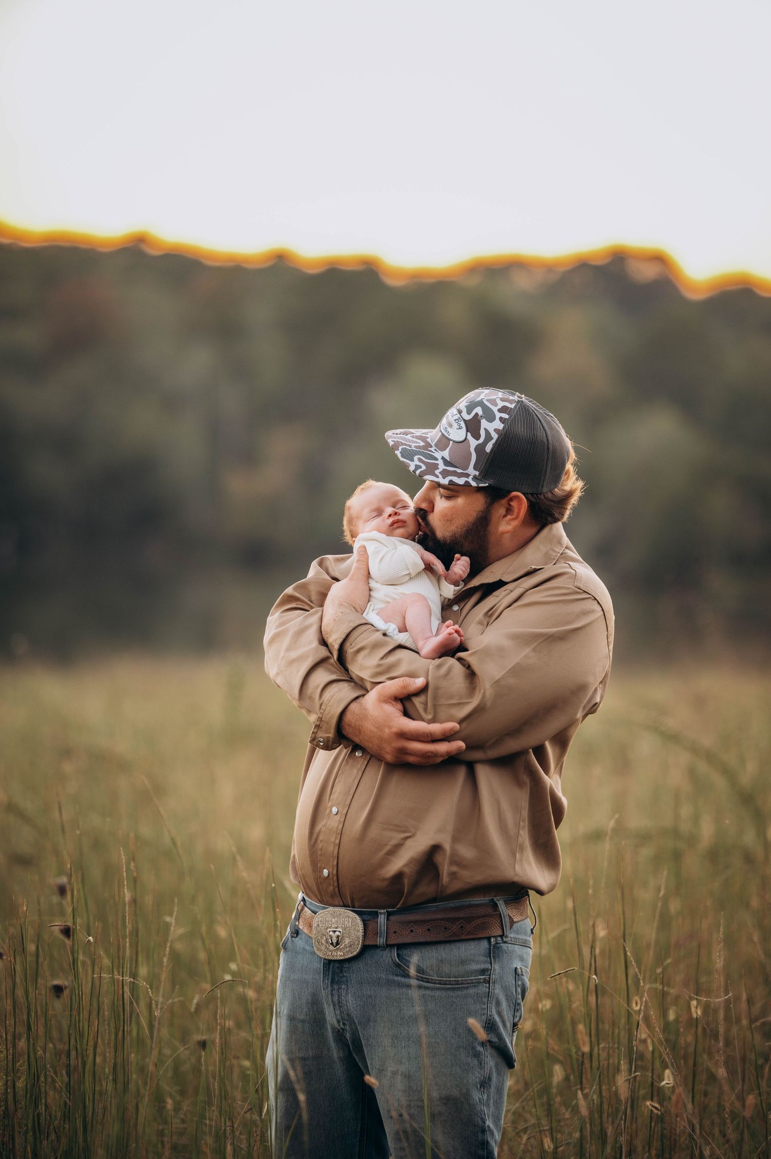 Parents embrace their newborn in a sunlit field at golden hour.