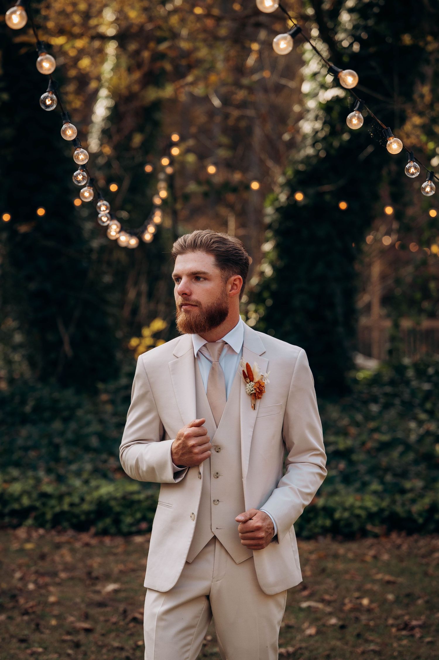 A groom in a light beige suit poses outdoors with autumn foliage and string lights in the background.