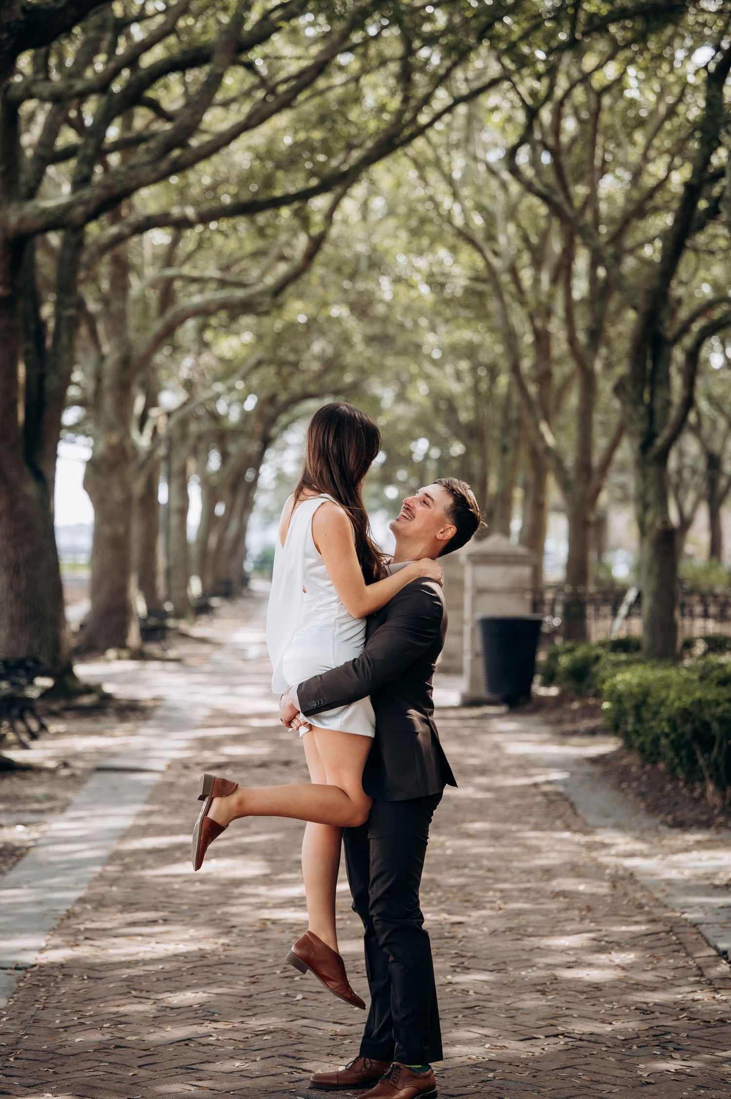 A couple embraces and spins joyfully under a canopy of oak trees along a tree-lined path.