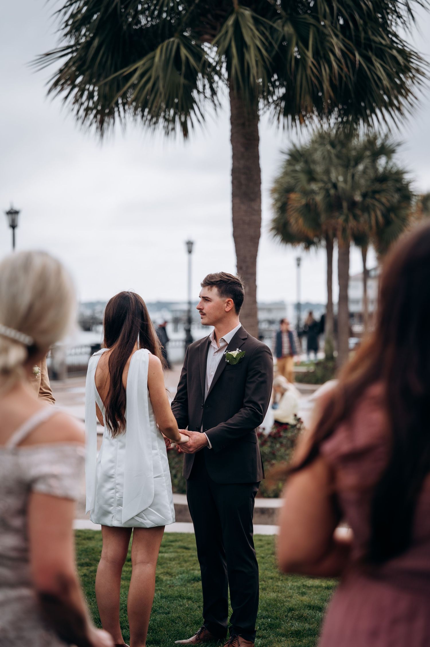 A romantic proposal scene unfolds on a grassy lawn with palm trees and onlookers in the background.