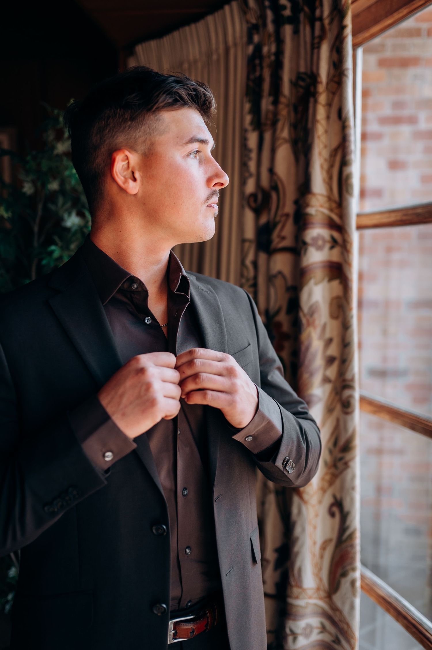 Groom buttoning his dark suit jacket while gazing out a window with ornate curtains.