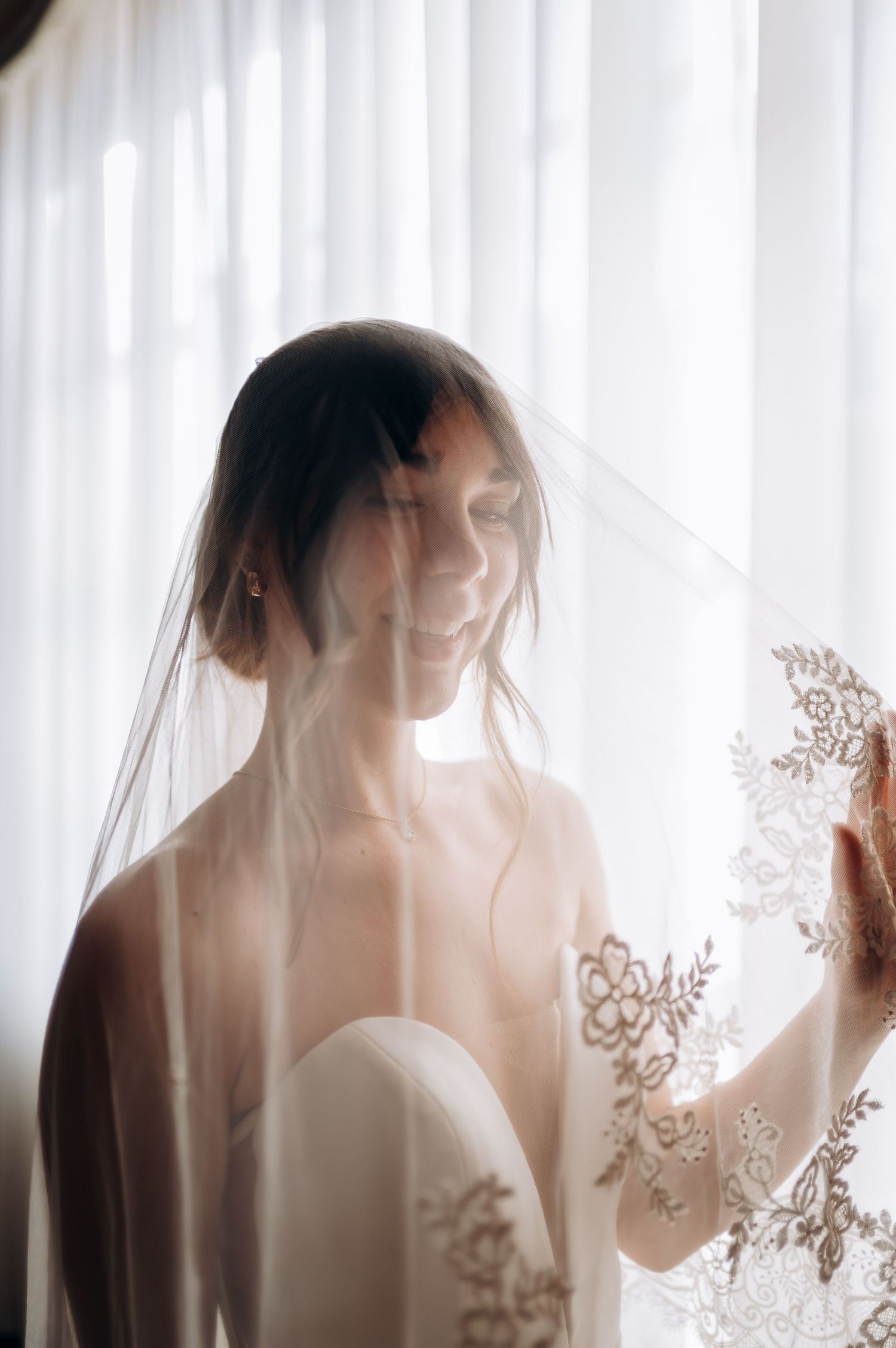Smiling bride draped in lace-trimmed veil stands before sheer curtains in soft natural light.