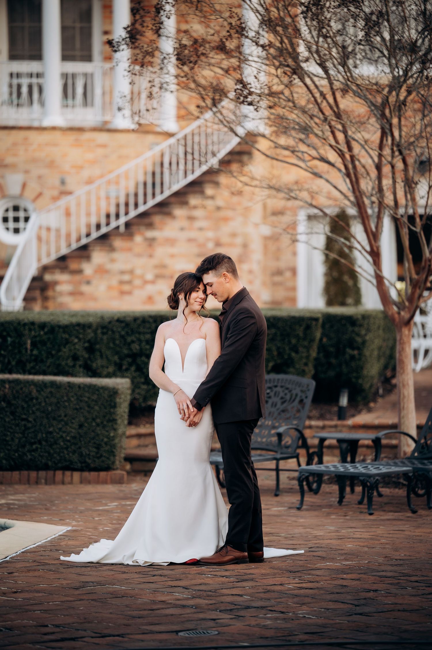 Bride and groom touch foreheads tenderly on brick patio outside elegant brick manor with white staircase.