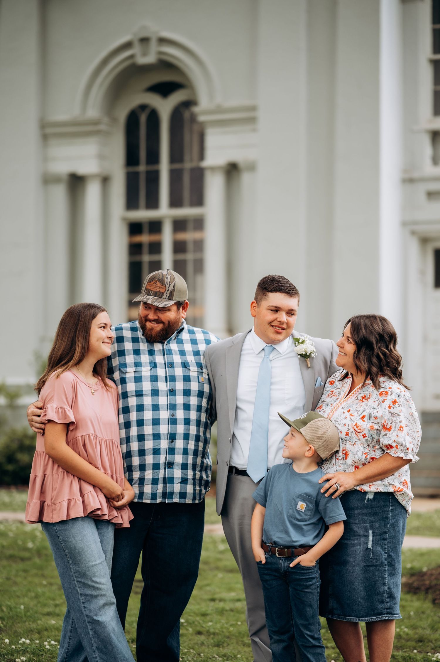 Family of five posing outdoors in front of a white church, dressed in casual and formal attire, smiling together.