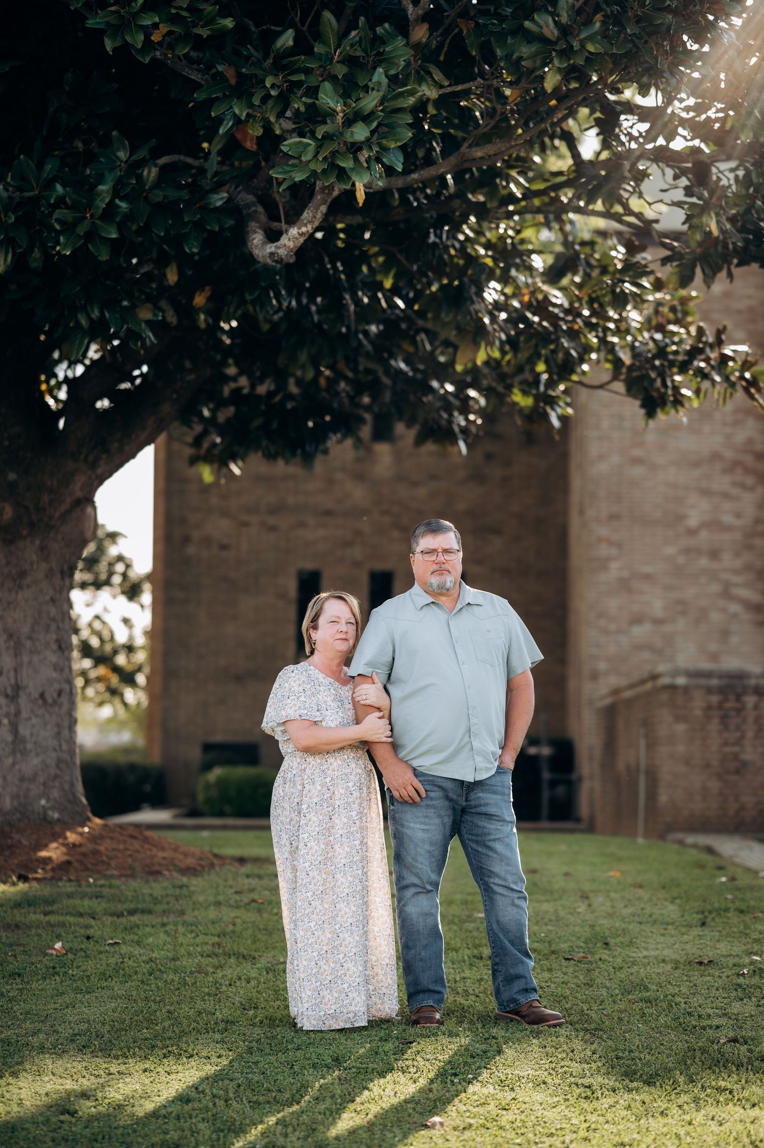 Couple standing together on green grass under a large tree outside a brick building in warm golden sunlight.