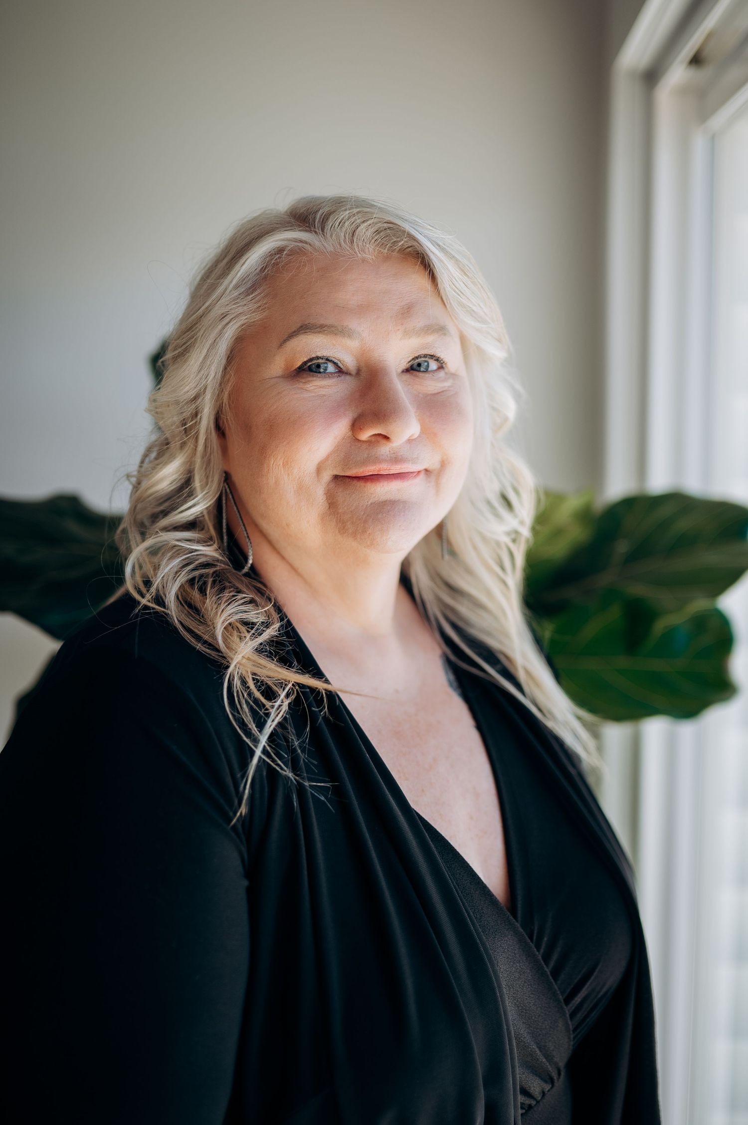 A smiling middle-aged woman with long silver-blonde hair wearing a black wrap top, posed near a window with green foliage.