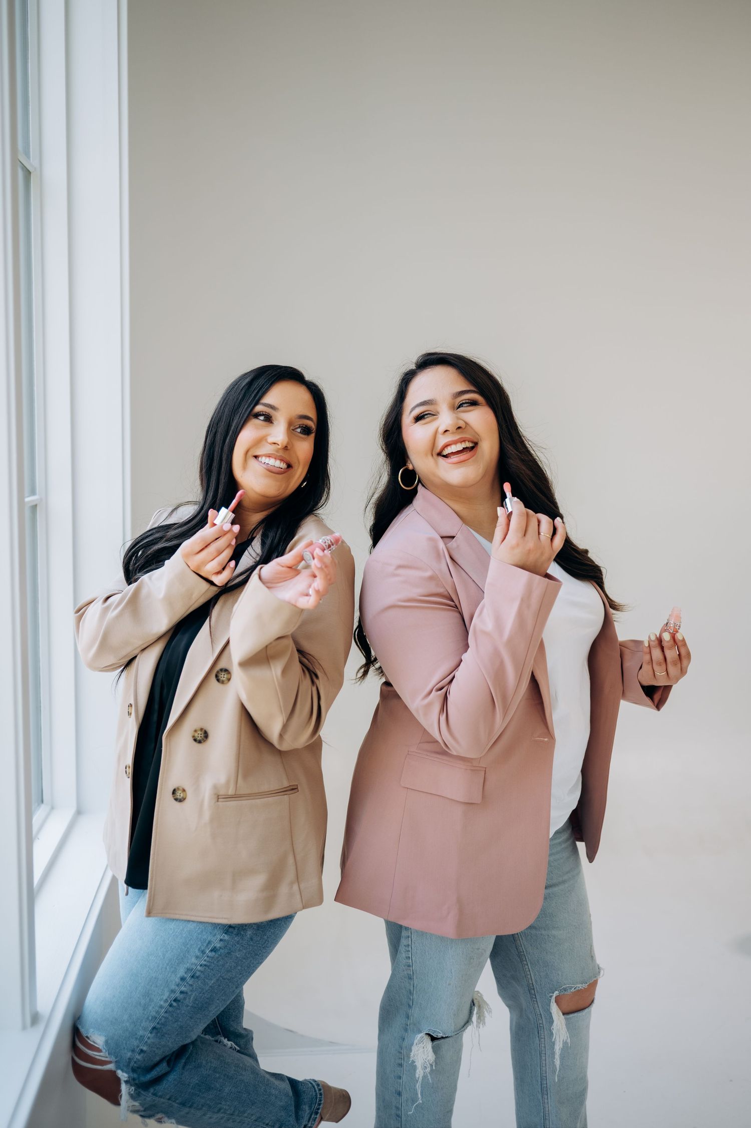 Two friends in stylish blazers and ripped jeans pose playfully by a window in a casual fashion photoshoot.