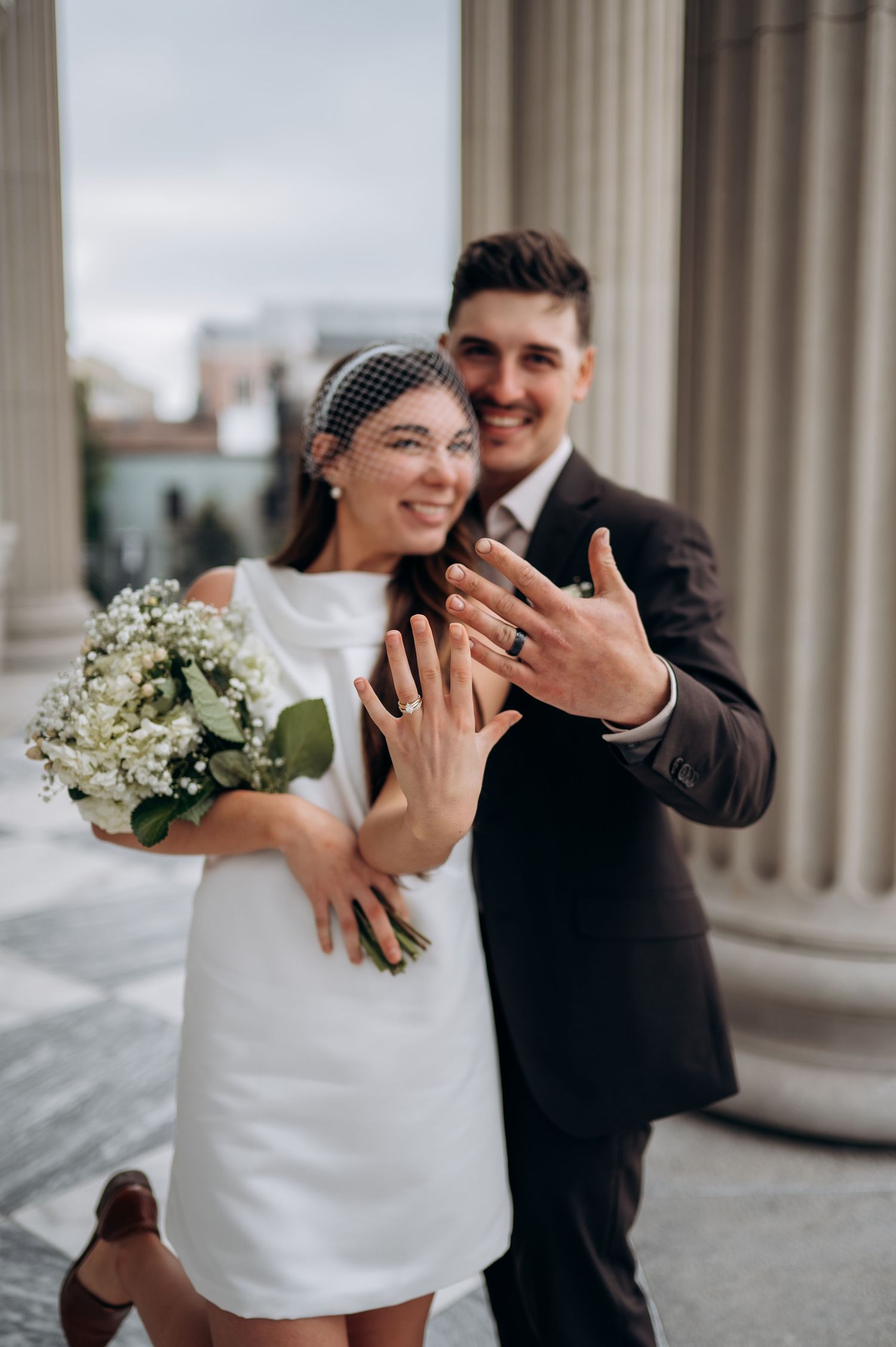 A joyful wedding couple embracing near classical columns with a white bouquet.