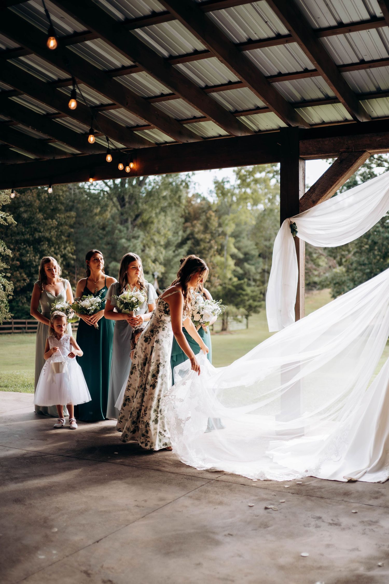 Bridesmaids help arrange a flowing white wedding veil in a rustic covered venue.