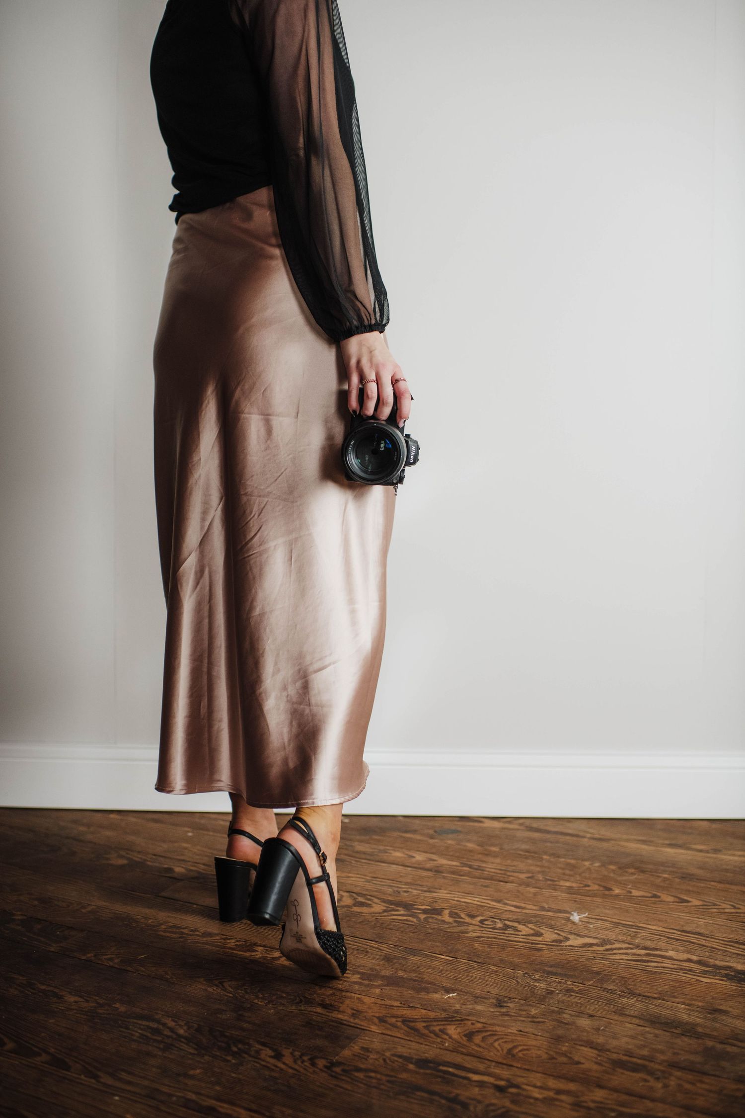 A person in a brown silk skirt and black sheer top stands on a wooden floor holding a camera.