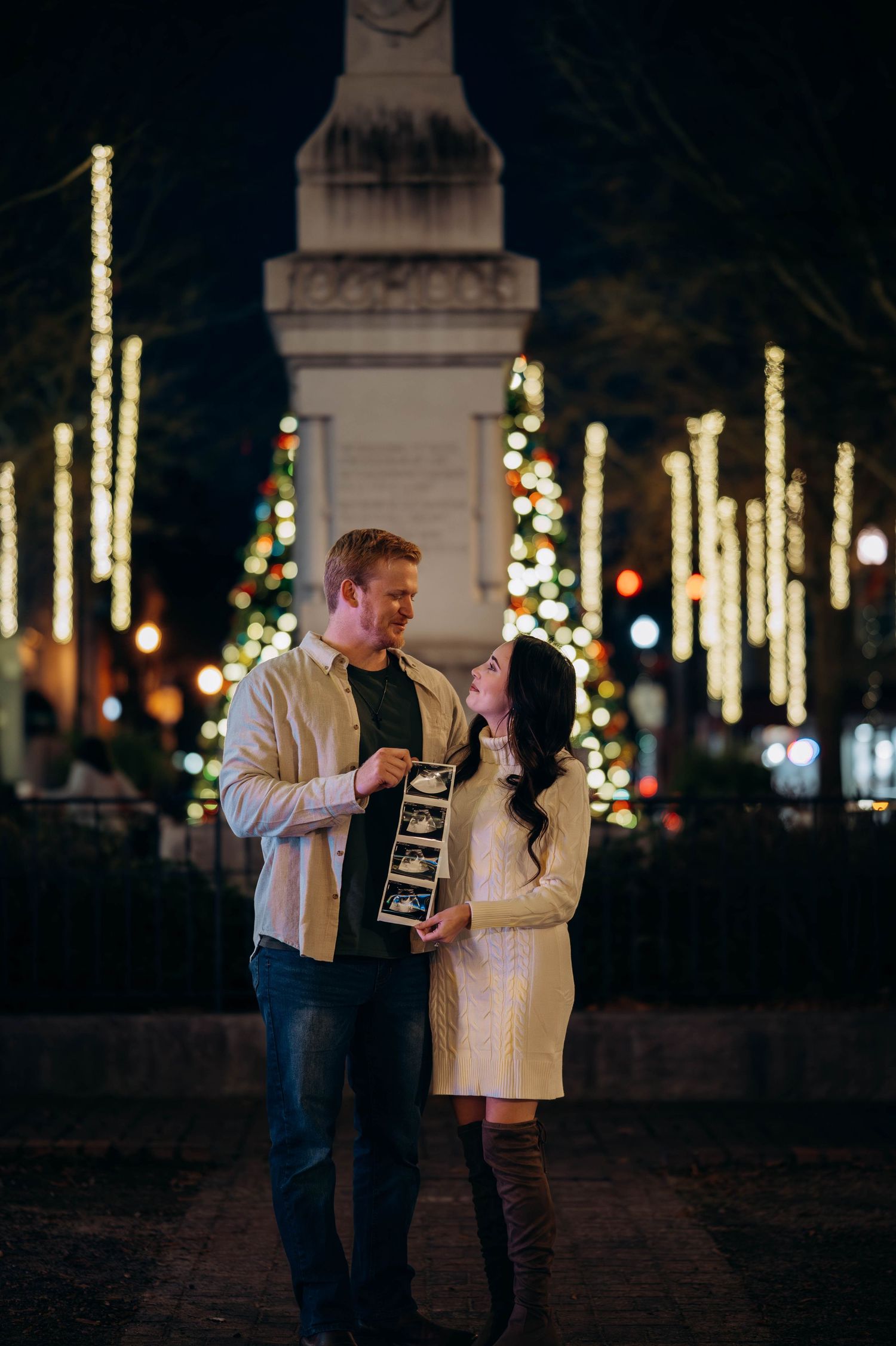 Couple holding ultrasound photos while standing among Christmas lights and decorated trees at night.