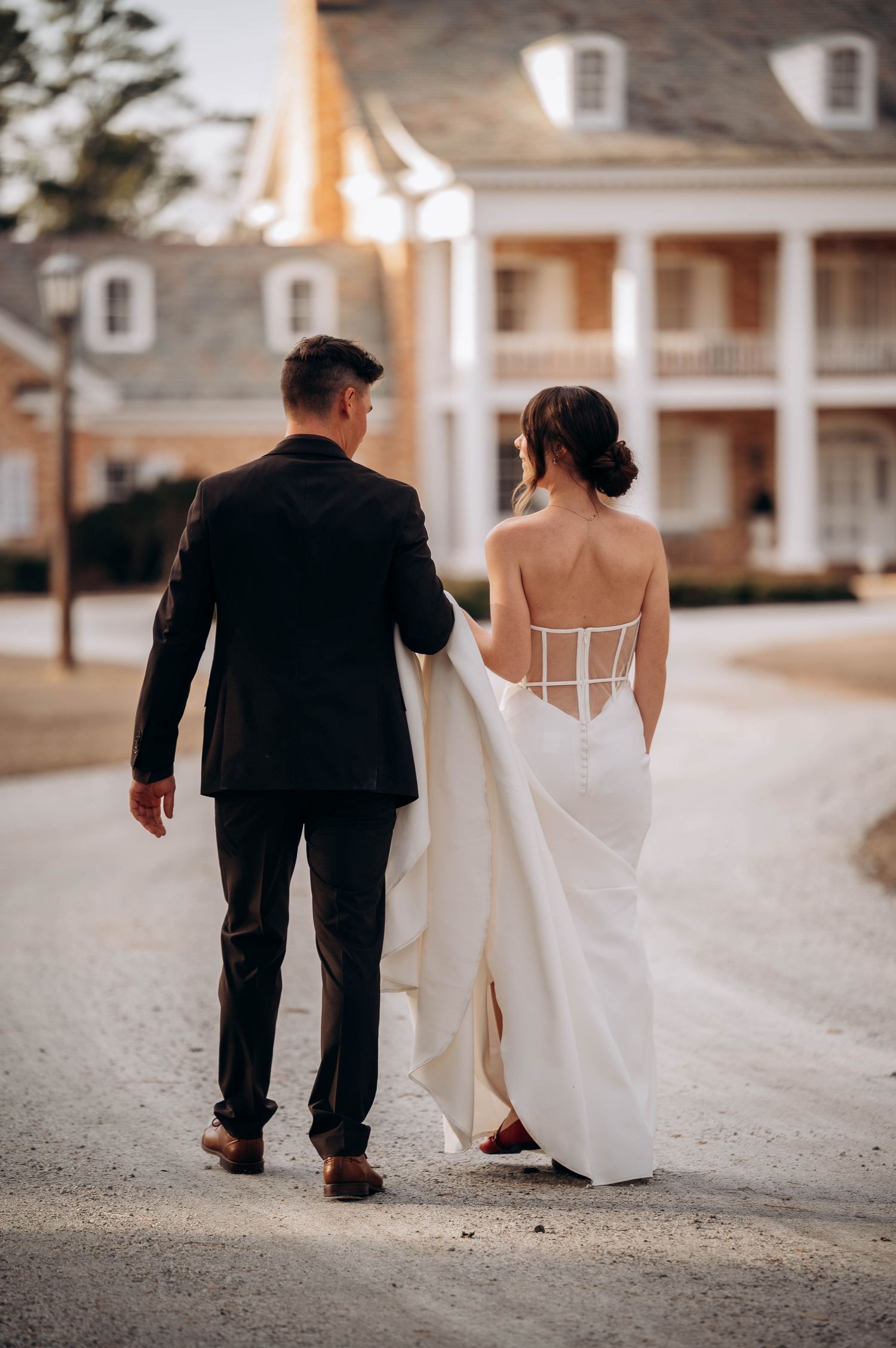 Bride in backless white gown and groom walk hand in hand toward a grand white colonial estate.
