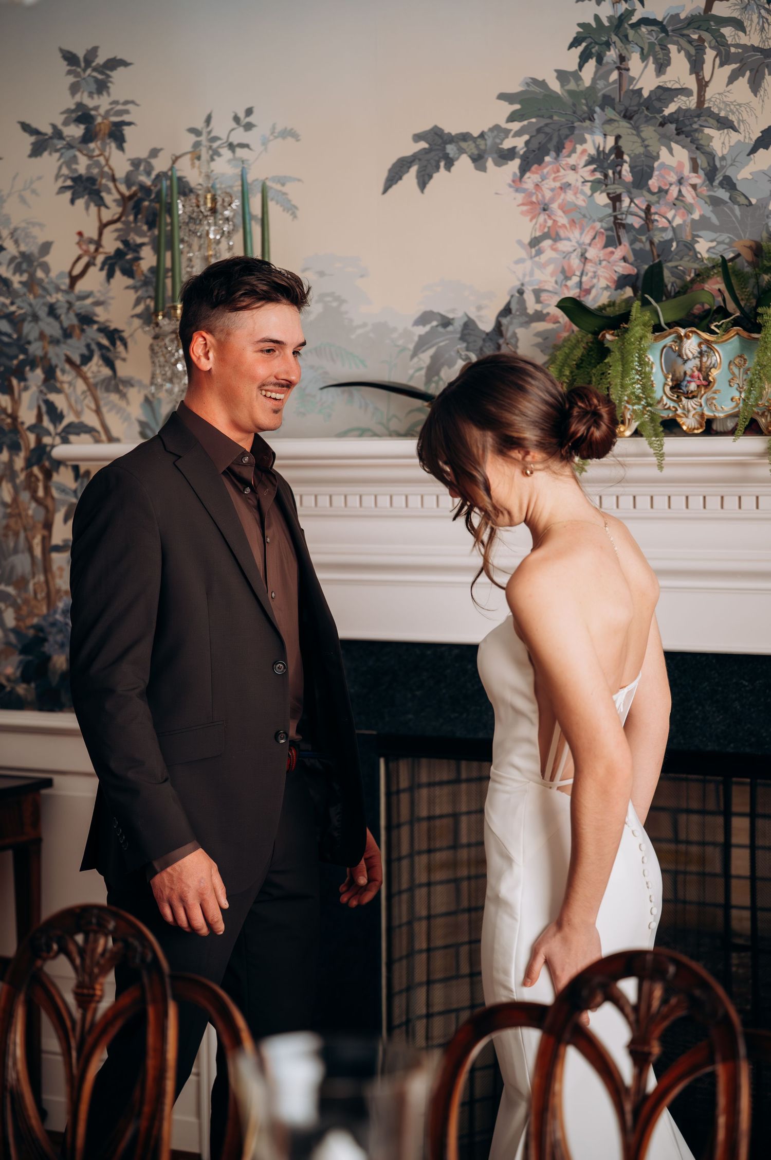 Groom in dark suit smiling at bride during first look moment beside ornate floral wallpaper fireplace.