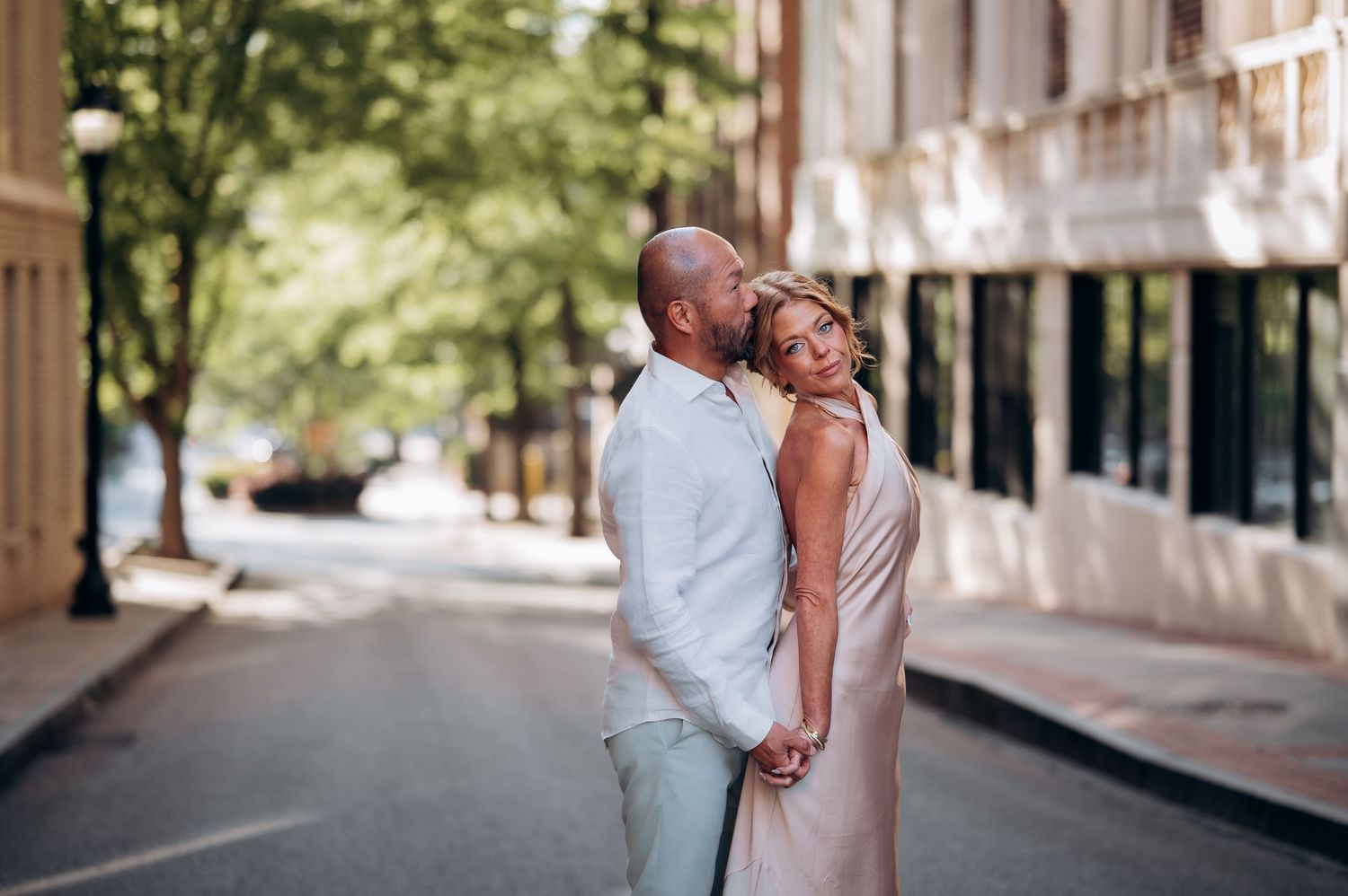 Romantic couple embracing on a quiet tree-lined urban street, woman in blush dress leaning back.
