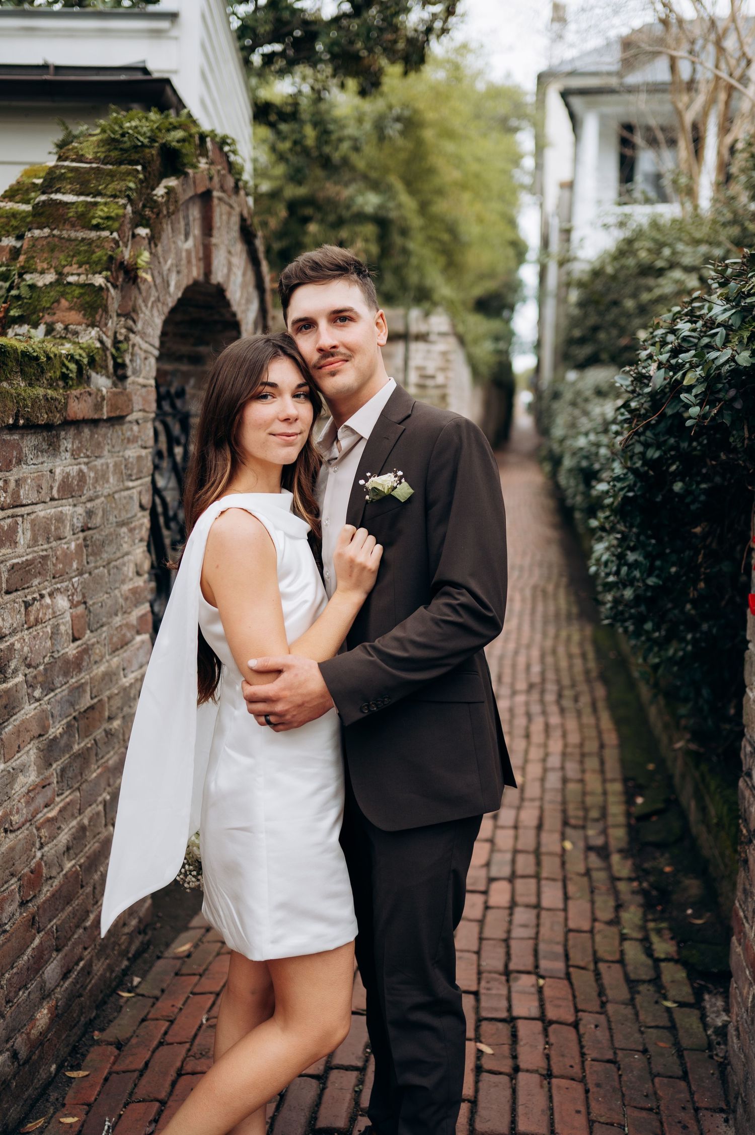 Bride and groom pose in charming brick alleyway, she wears a short white dress with cape detail.