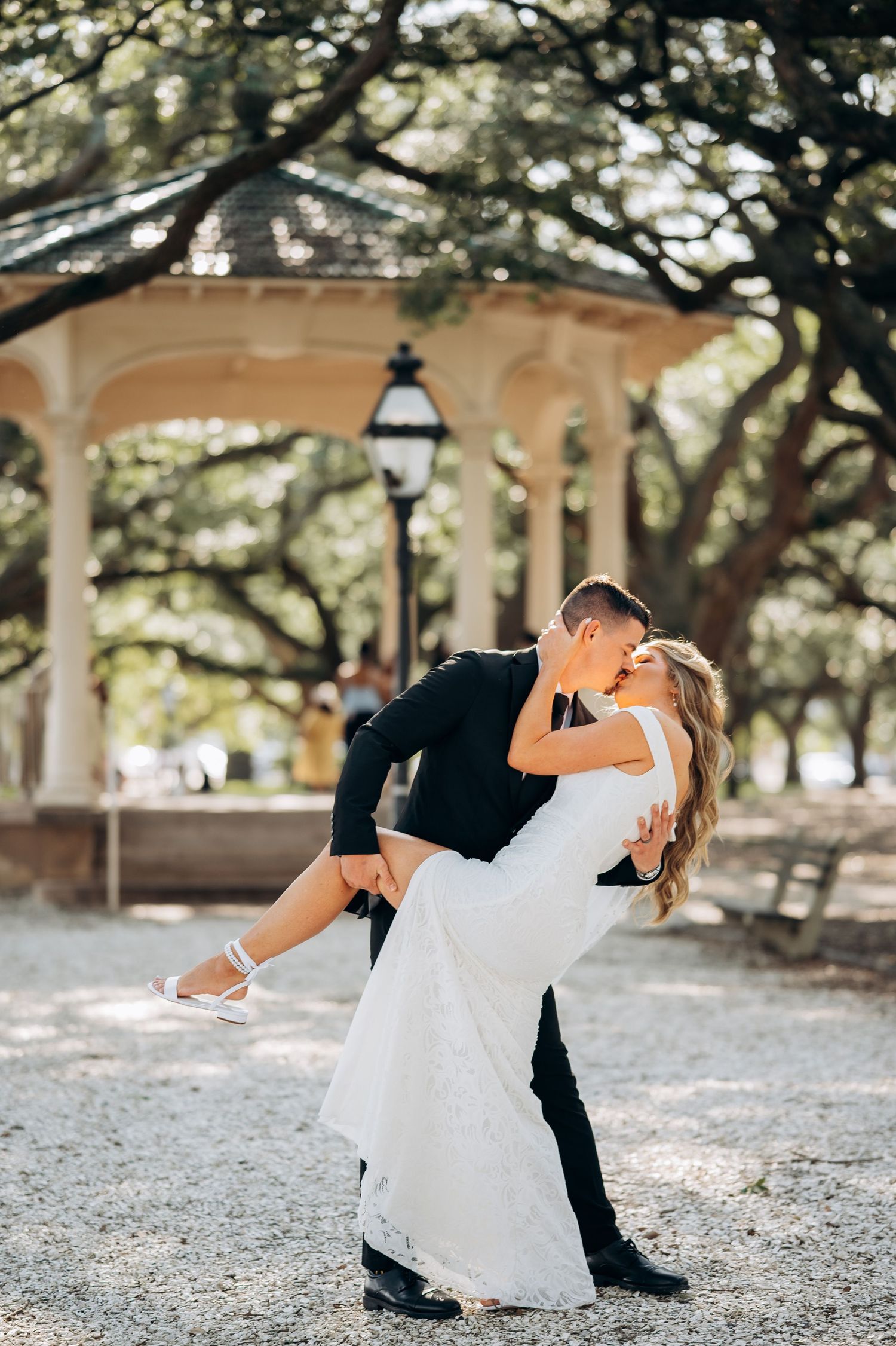 Groom dips and kisses bride in white lace gown near a gazebo surrounded by oak trees.