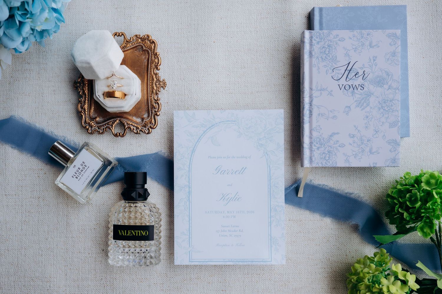 Elegant wedding flat lay with dusty blue invitation, Valentino perfume, ring box, and vow book on linen.