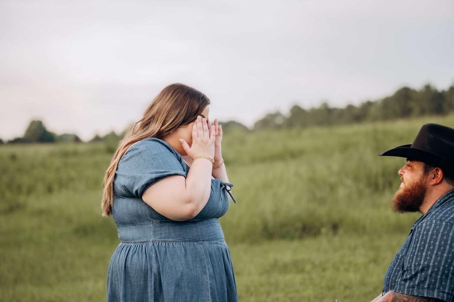 Overwhelmed woman covering her face with hands in joy during a proposal in a green field, bearded man in black hat nearby.
