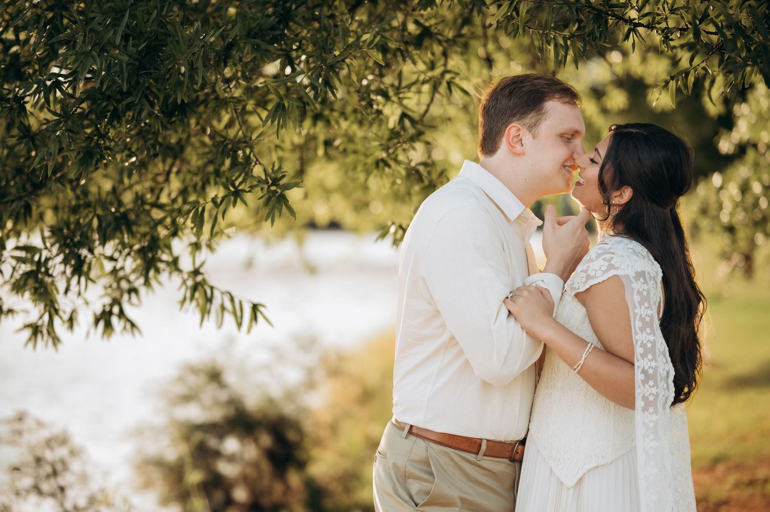 Couple embracing under willow tree branches near water in golden sunlight, wearing white wedding attire.
