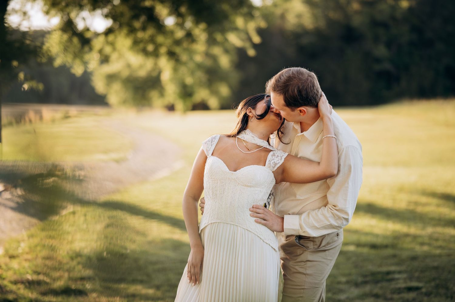 Couple sharing a tender moment in a sunlit green park, bride in white pleated dress with groom in khaki suit.