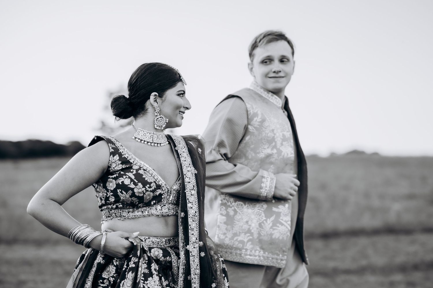 Black and white photo of a couple in traditional Indian wedding attire posing back-to-back in an open field.