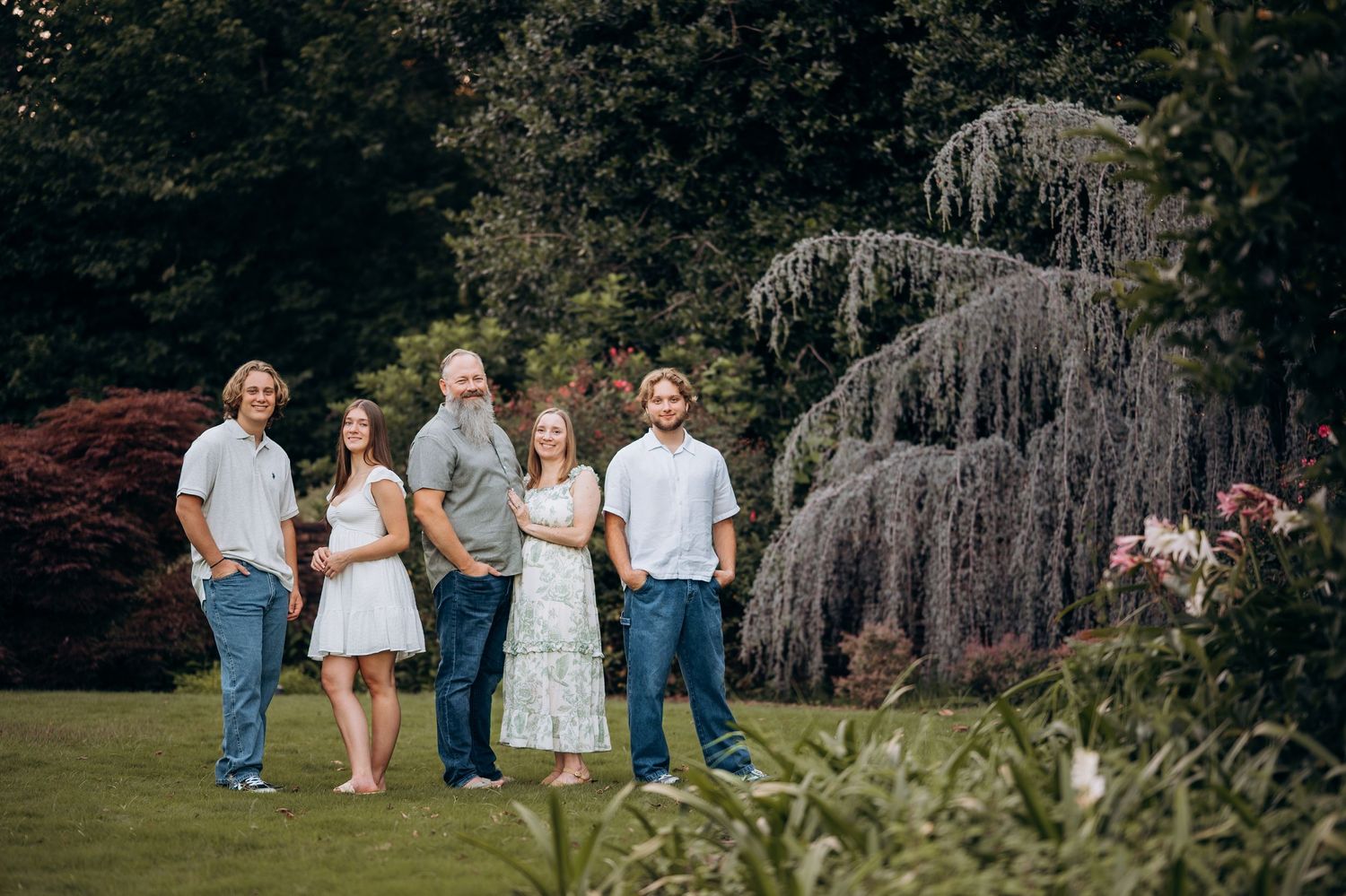 Family of six posing outdoors in a lush green garden with weeping trees and colorful foliage in the background.