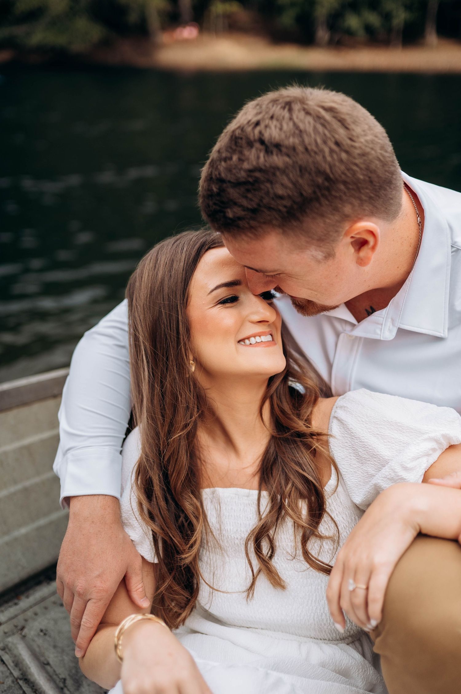 A couple shares an intimate moment while sitting by a lake in casual white attire.